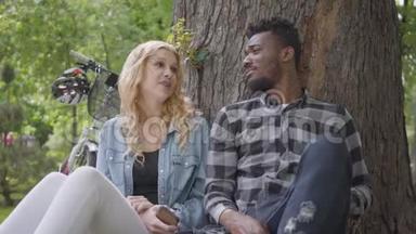 公园里坐在一棵老树下谈天说地的金发美女和美籍黑人帅哥